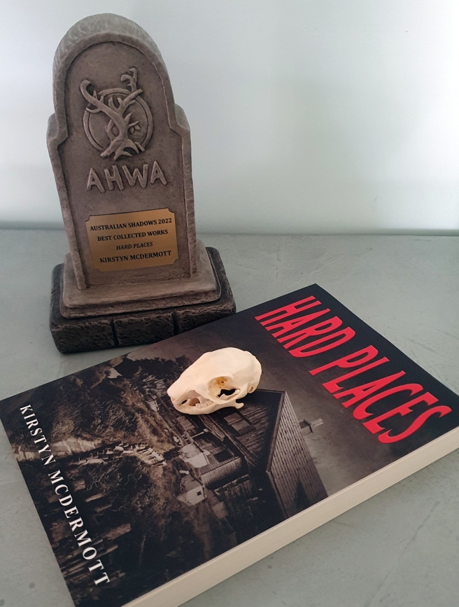 The trophy for the Australian Shadows Award 2022 for Best Collected Work, which is in the shape of a grey headstone with the AHWA haunted tree logo on the front. Alongside it sits a copy of the winning book, Hard Places by Kirstyn McDermott with a small squirrel skull on top. Because Halloween and why not?